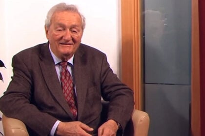 Egy idősebb úr ül egy világos székben, hátterében növényekkel és ajtóval. Az úr barna zakóban és piros mintás nyakkendőben van. A tekintete a kamera felé irányul és mosolyog.