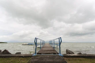 Egy fahíd vezet a Balaton viharos vizére, melynek hátterében felhős égbolt és a távoli hegyvonulatok láthatók. A kép előterében, a bal oldalon néhány nagyobb kődarab van, melyek a híd bejáratát szegélyezik. A híd kék fém korlátjai kiemelkednek a természetes színek között, hozzáadva egy modern hatást a klasszikus tájhoz.