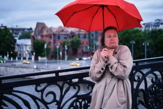 kabátos nő áll egy hídon piros esernyővel rossz időben