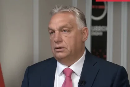 Orbán Viktor, Magyarország miniszterelnöke, sötét öltönyben és rózsaszín nyakkendőben interjút ad, komoly arckifejezéssel, a háttérben egy magyar zászlóval