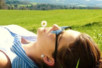 Fiatal nő hanyatt fekve élvezi a tavaszi napsütést egy smaragdzöld, friss fűvel borított mezőn. Csíkos, kék-fehér felsőt visel, napszemüveg takarja a szemét, fülében gyöngy fülbevaló csillog. Ajkai között fehér margarétavirágot tart, miközben láthatóan pihen és kiélvezi a meleg időjárást. A háttérben távolabb zöld dombvidék és erdős táj húzódik, jelezve az idilli vidéki környezetet és a beköszöntő tavaszi időszakot. A kép tökéletesen illusztrálja a természet ébredését és a szabadtéri kikapcsolódás örömét a tavasz első meleg napjain.