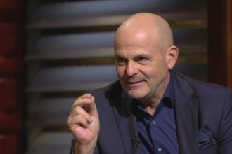 Moldován András öltönyben haj nélkül mutogat egy stúdióban