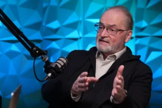 Farkas András NyugdíjGuru a hvg.hu stúdiójában magyaráz a nyugdíjakról kék geometrikus háttér előtt, mikrofonba beszélve.