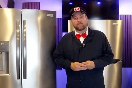 Egy piros csokornyakkendős, szakállas, magas férfi munkaruhában és baseballsapkában beszél a kamerába három hűtő előtt.