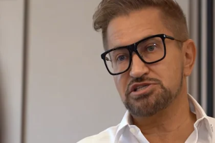 Jákob Zoltán fekete keretes szemüvegben, szőke, balra fésült hajjal, szakállasan, fehér ingben beszél.