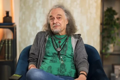 Laár András megrendült tekintettel, félhosszú hajjal, zöld pólóban, szürke kapucnis, cipzáros pulcsiban és farmerben egy kék kanapén ül, miközben a kamerába néz.