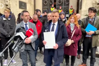 Magyar Péter kék kabátban, zakóban és nyakkendőben, fehér ingben, a csapatával a Legfőbb Ügyészség és a Kúria épülete előtt áll, miközben a kezében egy dossziét tart, és mikrofon előtt beszél.