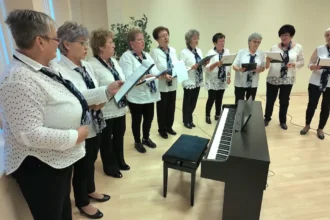 Nyugdíjas nők fehér felsőben, sötét sálban egy zongora előtt, kottát tartva, félkörben állva énekelnek.