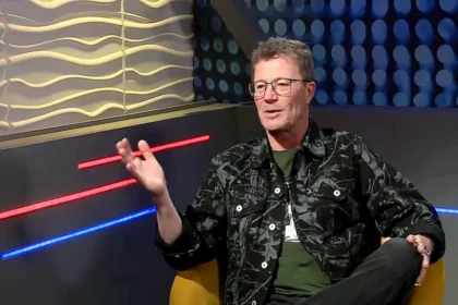 Stohl András egy mustársárga székben ülve, fekete, mintás ingben, szemüvegben, gesztikulálva beszél.