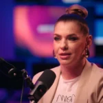 Dér Heni barack színű blézerben, fülbevalóban, kontyba fogott hajban, sminkben mikrofonba beszél, miközben kamerába néz.