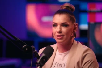 Dér Heni barack színű blézerben, fülbevalóban, kontyba fogott hajban, sminkben mikrofonba beszél, miközben kamerába néz.