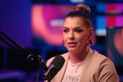 Dér Heni barack színű blézerben, fülbevalóban, kontyba fogott hajban, sminkben mikrofonba beszél, miközben kamerába néz.