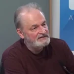 Farkas András vörös pólóban, ősz szakállal és hajjal, kopaszodva, jobbra nézve a mikrofonba beszél.