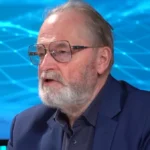 Farkas András kék zakóban, sötétkék ingben, szemüvegben, szakállasan, balra nézve beszél egy kék grafika előtt.