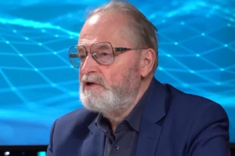 Farkas András kék zakóban, sötétkék ingben, szemüvegben, szakállasan, balra nézve beszél egy kék grafika előtt.