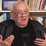 Havas Henrik fekete pólóban, fekete bőrdzsekiben, szemüvegben, ősz hajjal, indulatosan beszél a mikrofonba egy könyvespolc előtt.