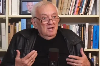 Havas Henrik fekete pólóban, fekete bőrdzsekiben, szemüvegben, ősz hajjal, indulatosan beszél a mikrofonba egy könyvespolc előtt.