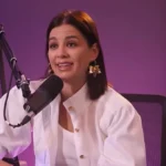 Ördög Nóra fehér kabátkában, fehér pólóban, barna hajjal, arany színű fülbevalóban, ciklámen színű háttér előtt nyilatkozik, miközben egy széken ülve a mikrofonba beszél.