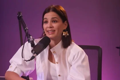 Ördög Nóra fehér kabátkában, fehér pólóban, barna hajjal, arany színű fülbevalóban, ciklámen színű háttér előtt nyilatkozik, miközben egy széken ülve a mikrofonba beszél.