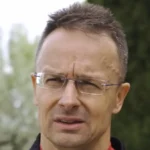 Szijjártó Péter szemüvegben, kócos hajjal, zöld háttérrel, sportruhában beszél, miközben gondterhelten néz balra.