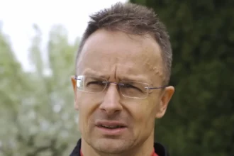 Szijjártó Péter szemüvegben, kócos hajjal, zöld háttérrel, sportruhában beszél, miközben gondterhelten néz balra.