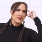 Tóth Gabi fekete felsőben, befont barna hajjal, erős sminkben enyhén balra és felfelé néz fehér háttér előtt.