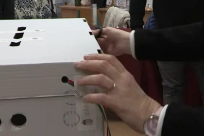 Egy szavazóurnát bont fel a választási bizottság női tagja fekete blézerben és fehér ingben, gyűrűben és karórában.