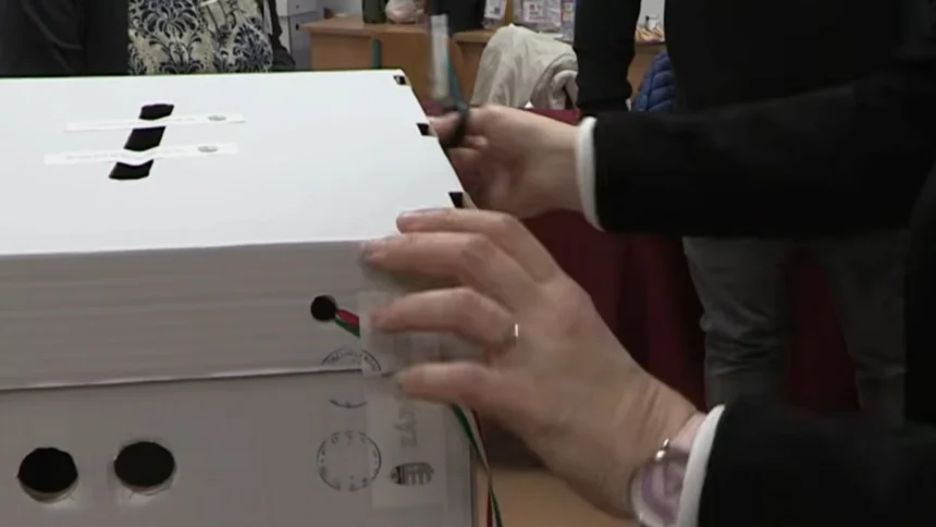 Egy szavazóurnát bont fel a választási bizottság női tagja fekete blézerben és fehér ingben, gyűrűben és karórában.