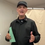 Vujity Tvrtko fekete ingben és sapkában, zöld szavazólapot a kezében tartva beszél egy szavazófülke előtt.