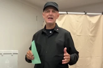 Vujity Tvrtko fekete ingben és sapkában, zöld szavazólapot a kezében tartva beszél egy szavazófülke előtt.